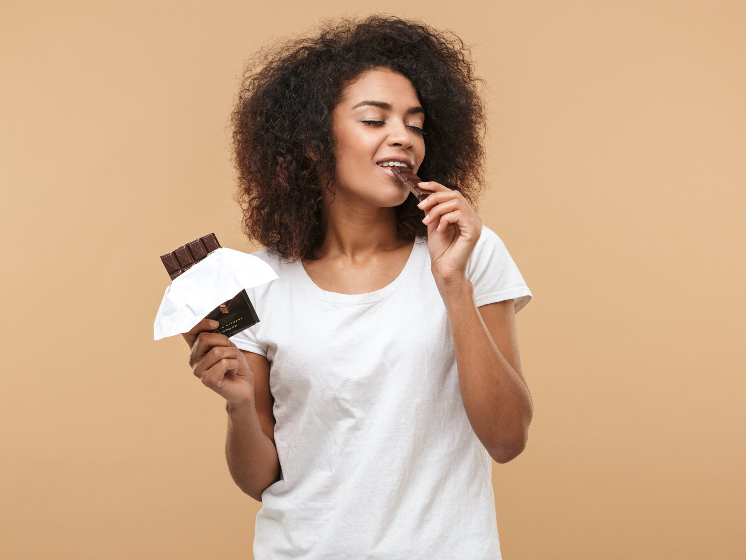 Portrait of a smiling young african woman eating chocolate bar isolated over beige background